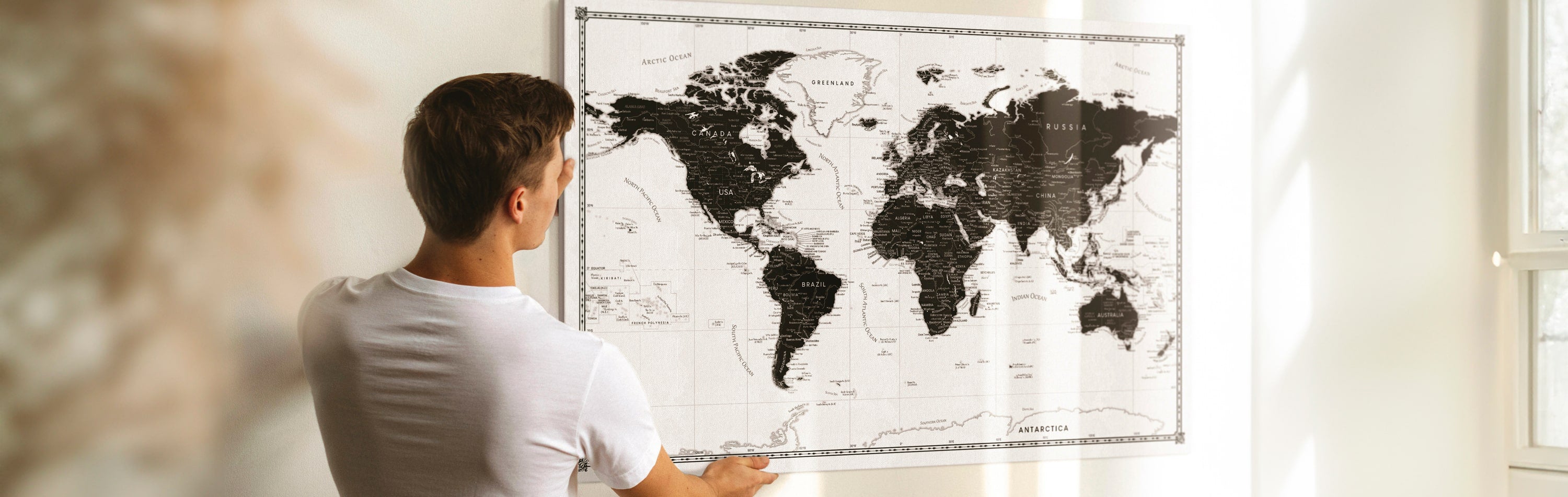 A man contemplating a black and white world map pin board on a wall, highlighting destinations using golden push pins - an elegant world map wall art for travelers, available on canvas, perfect for tracking and planning global journeys.