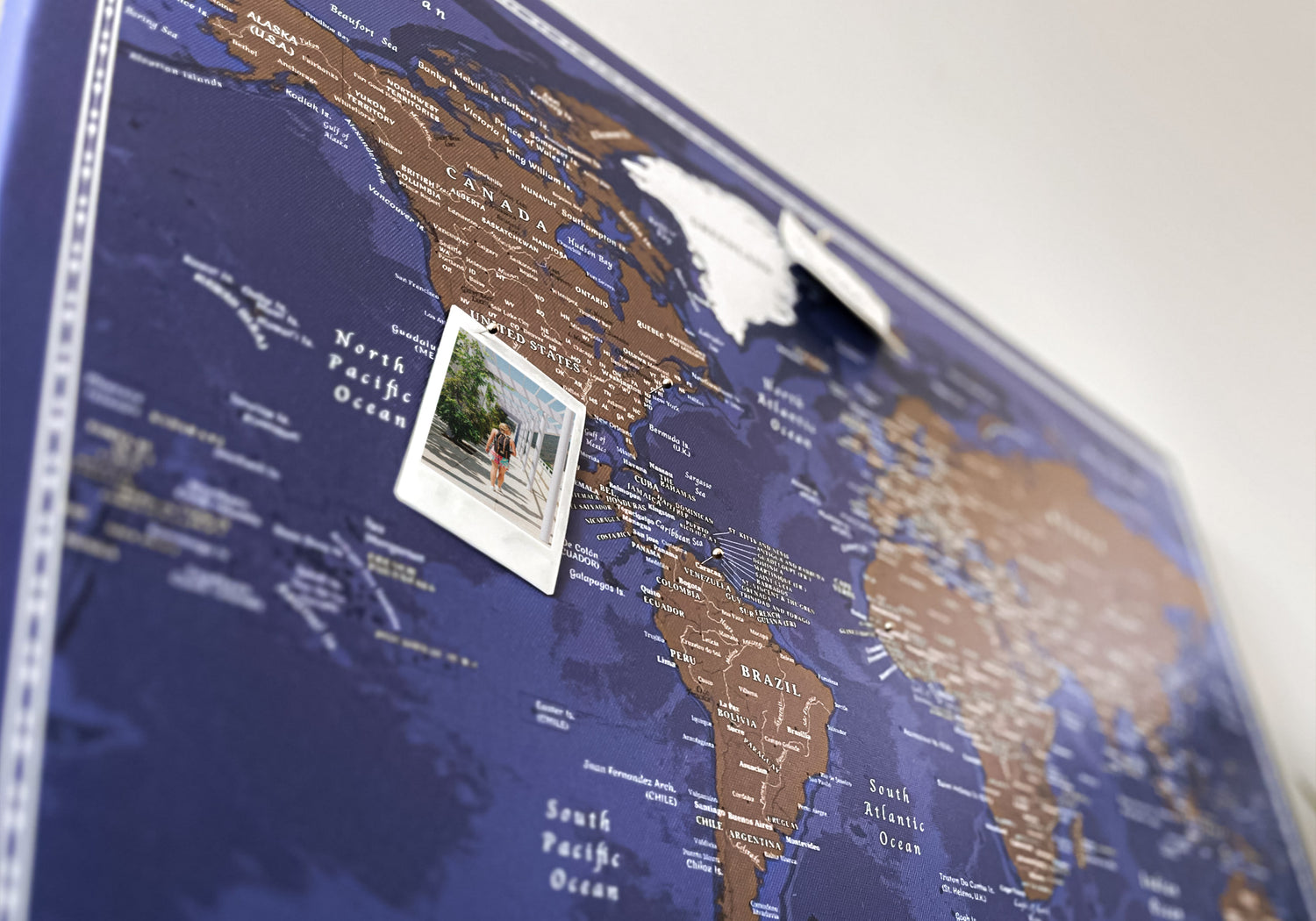 Diagonal view of a blue and brown push pin world map pinboard highlighting North America, an ideal travel map for marking destinations with a pinned polaroid, perfect as world map wall art for travel enthusiasts.